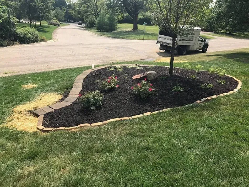 Mulch flower bed with red blooms and a signpost beside a driveway in a grassy yard