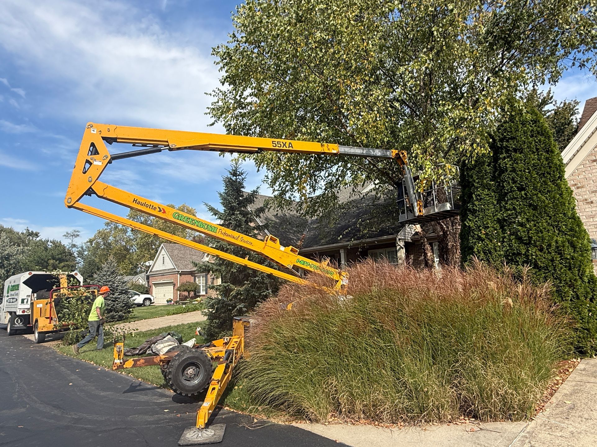 Yellow boom lift trimming overgrown bushes beside a suburban road