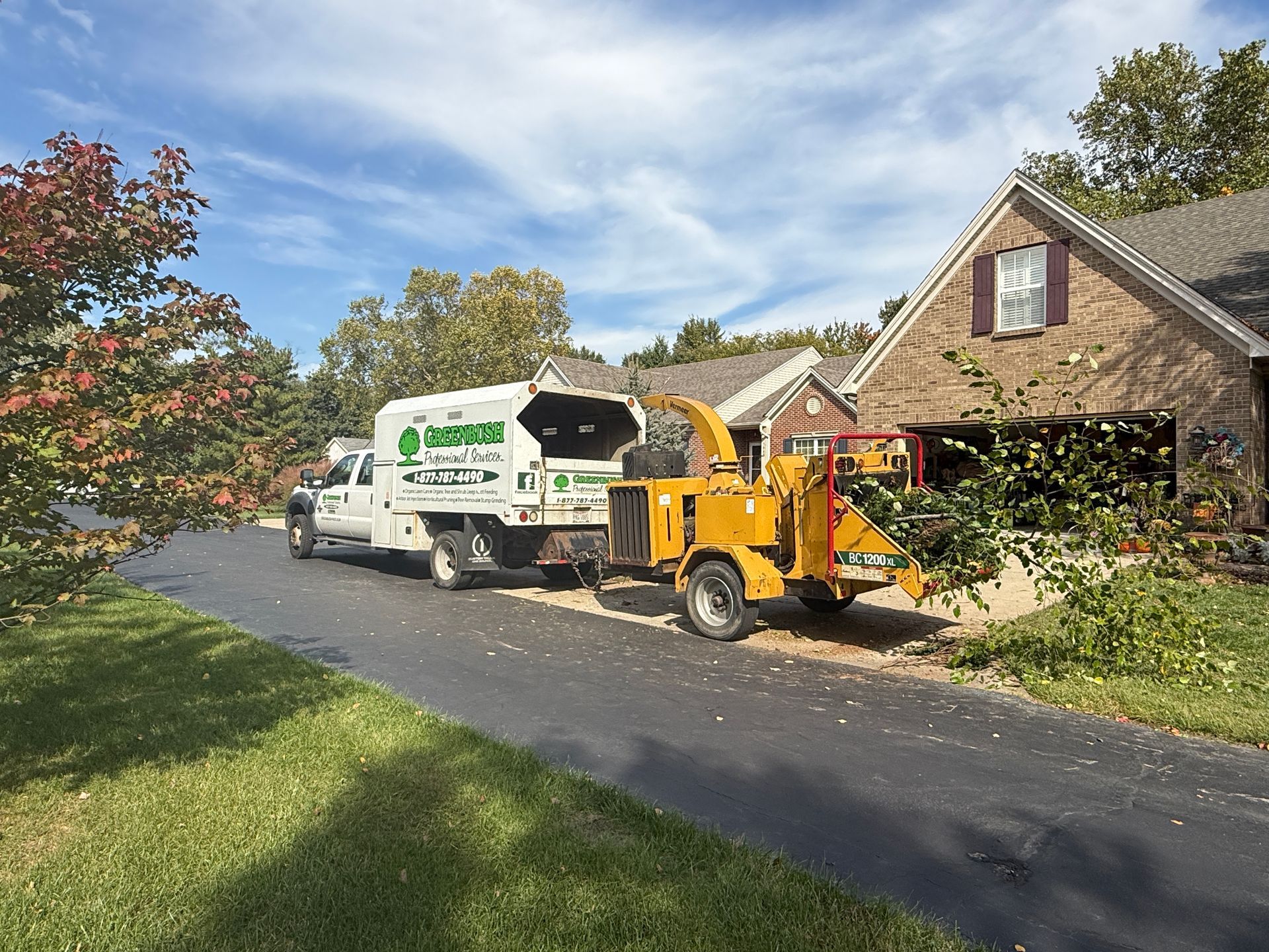 Landscaping truck and wood chipper on a driveway beside a brick house and autumn trees