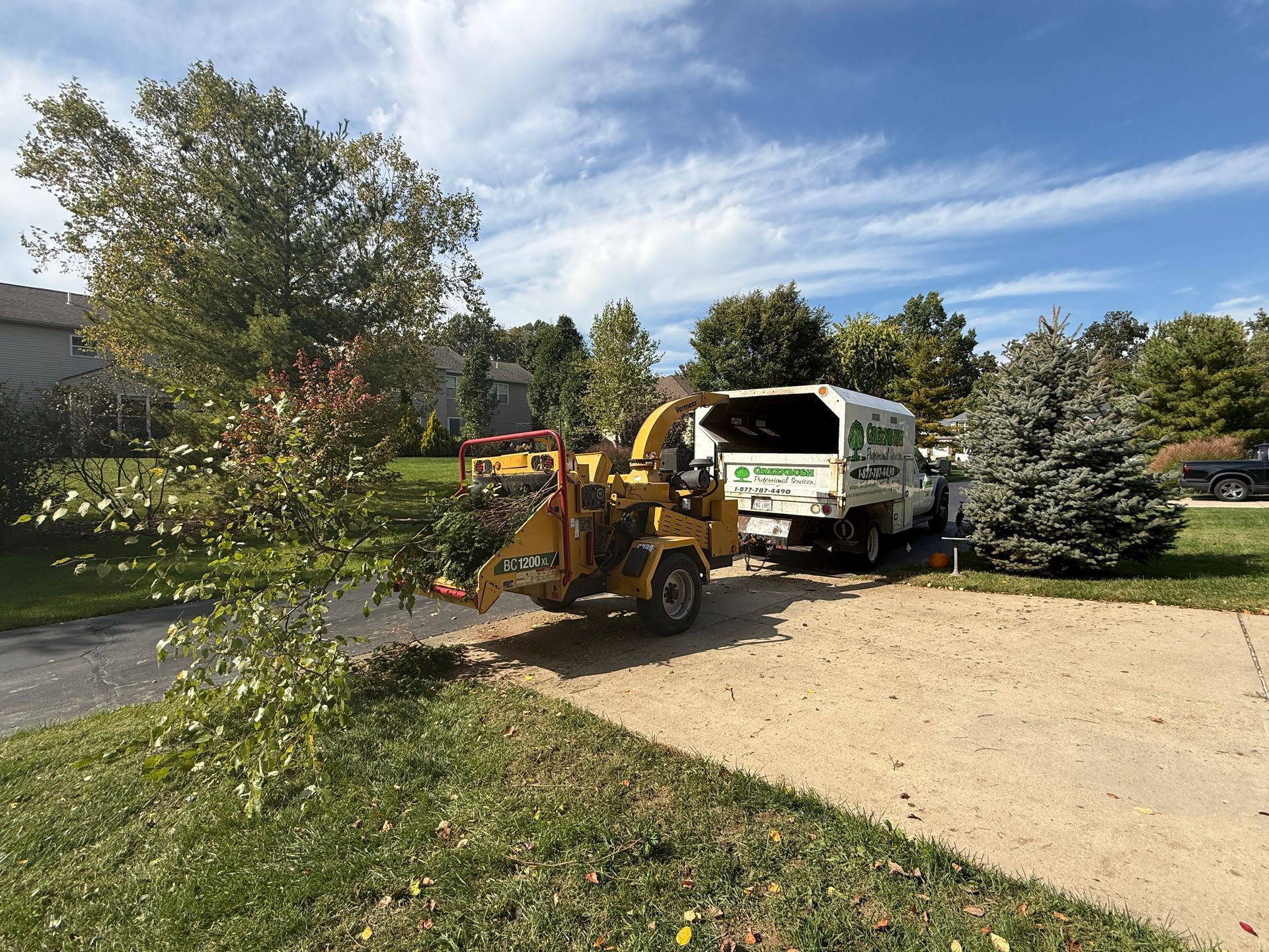 Yellow loader with raised bucket beside a white truck on a driveway under a partly cloudy sky
