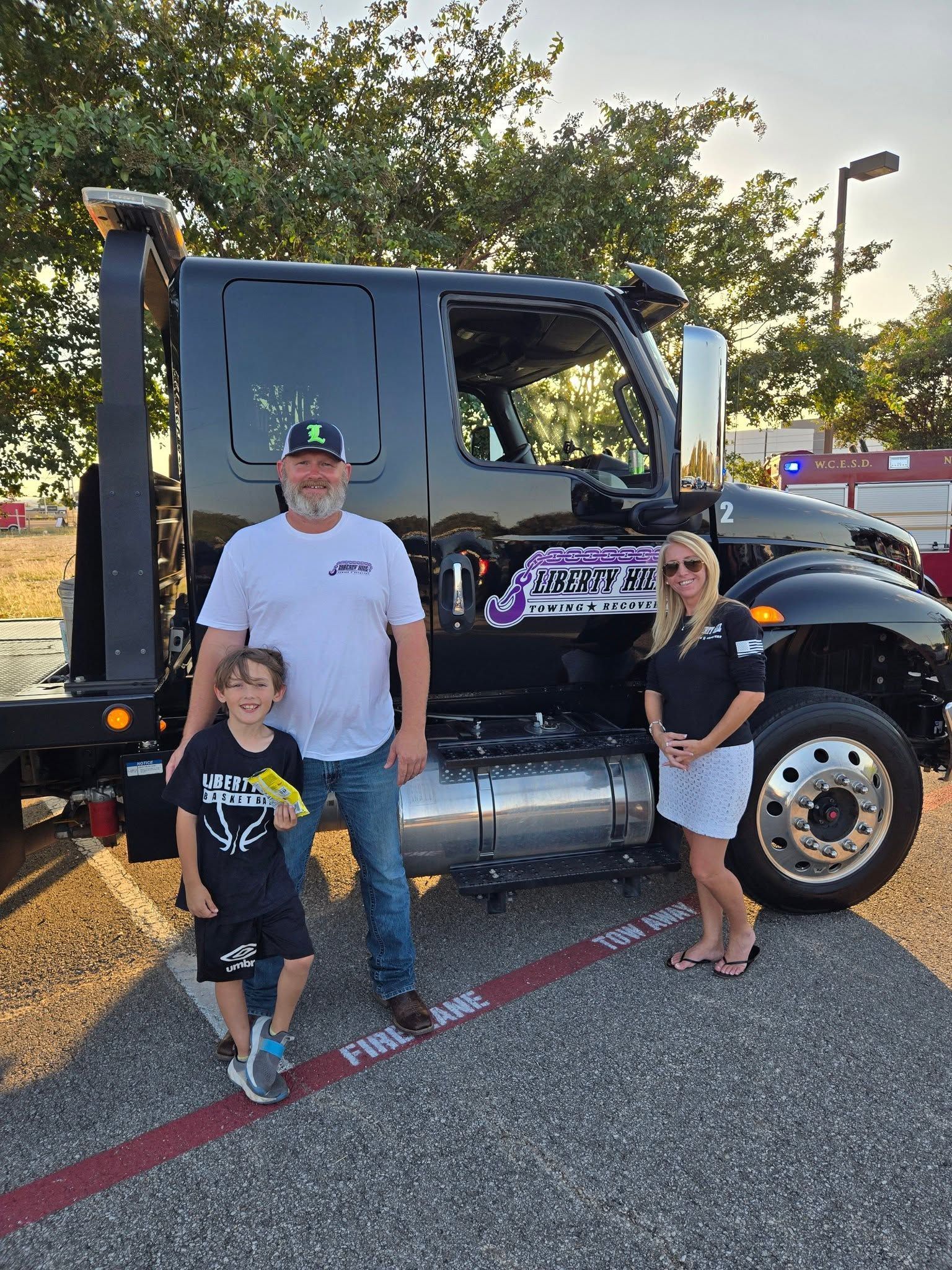 Three people stand beside a black truck with a purple logo in a parking lot at sunset.