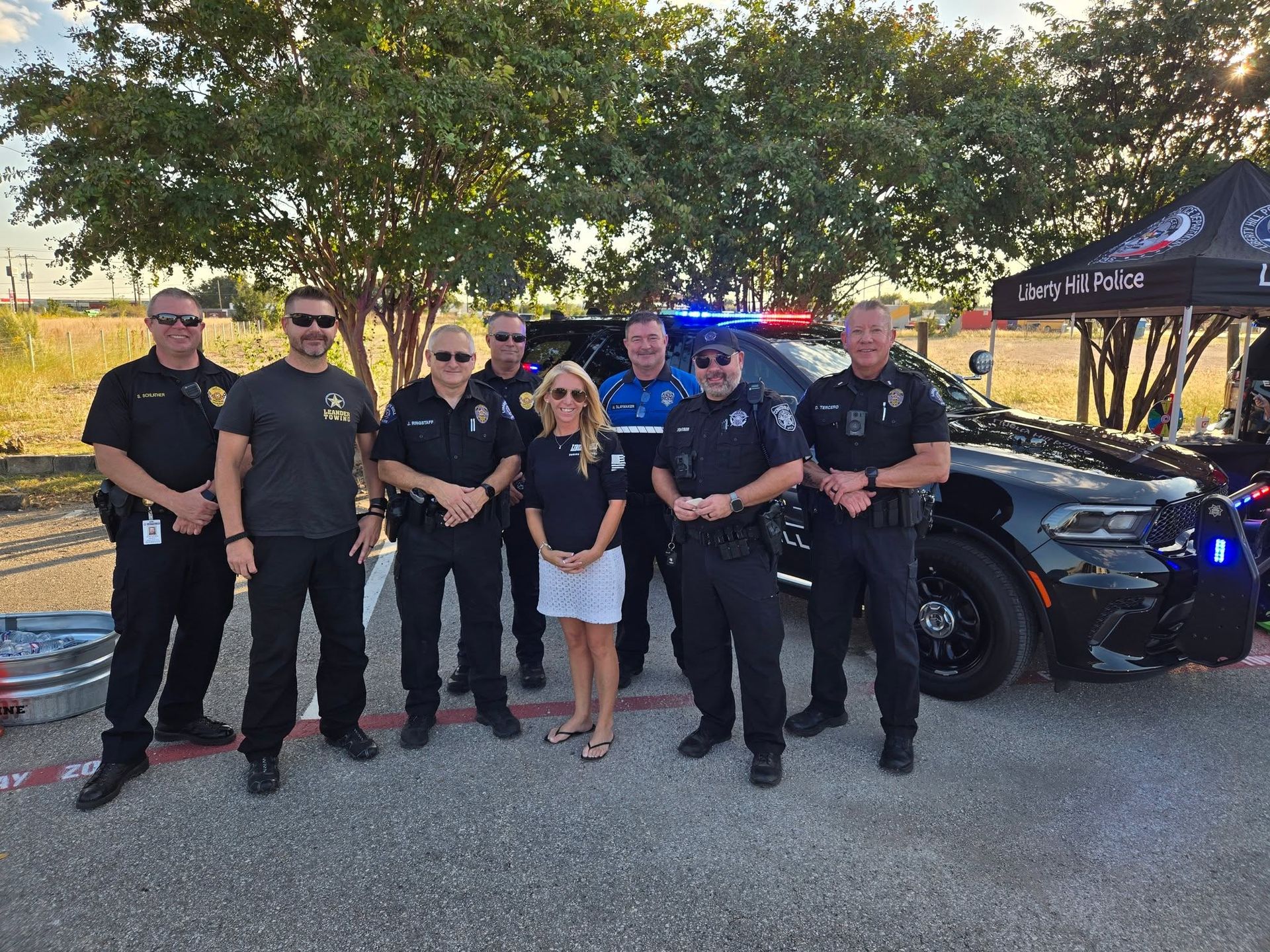 Police officers and a child pose beside a black police car at an outdoor event.