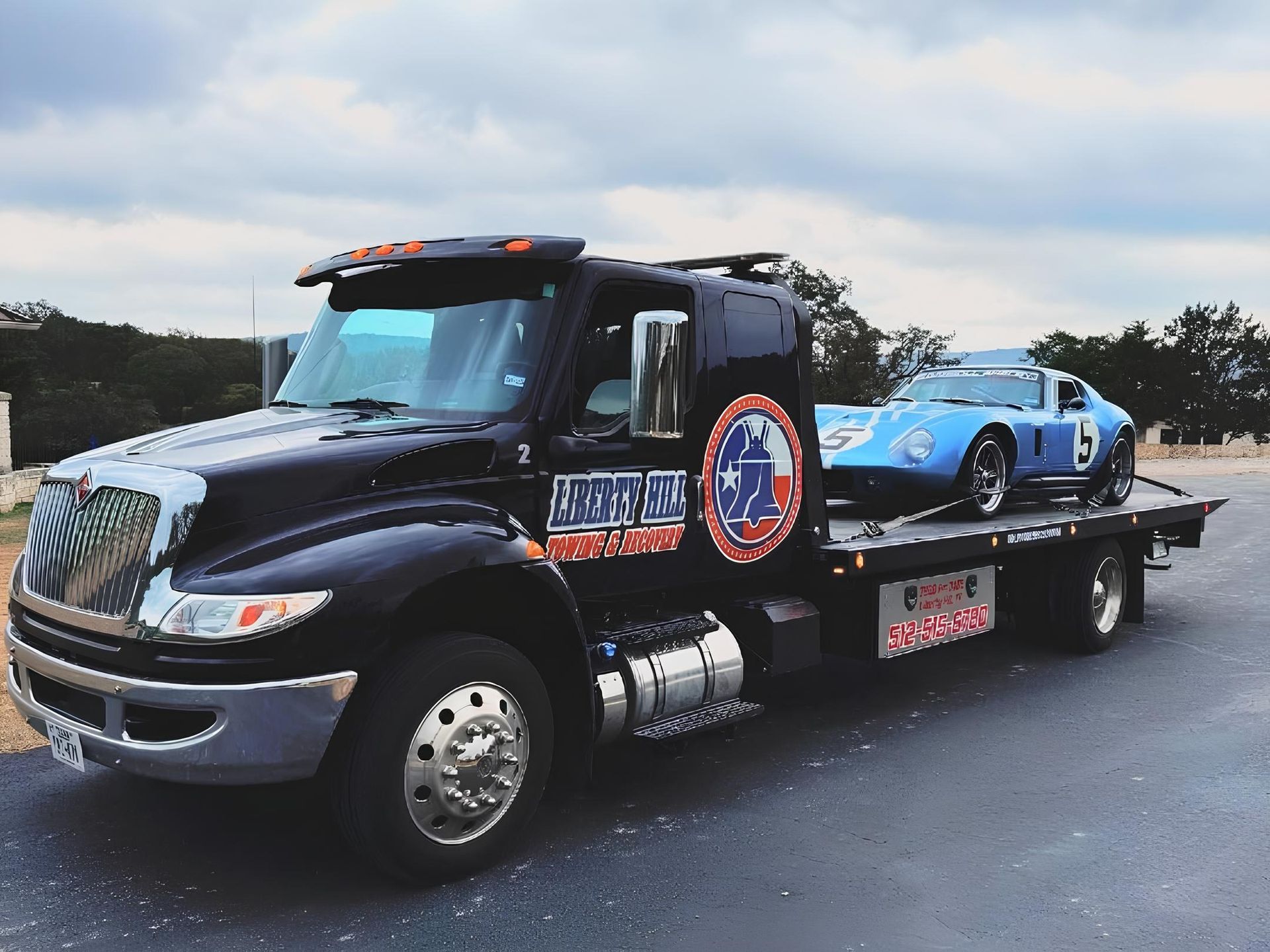 Black tow truck carrying a blue race car on a flatbed, parked on a road under cloudy sky