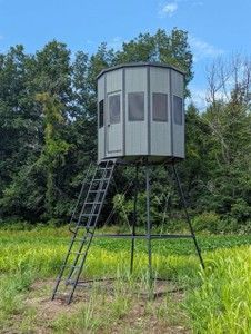 Elevated hunting blind in a field with ladder access and surrounding trees. Gray and black.