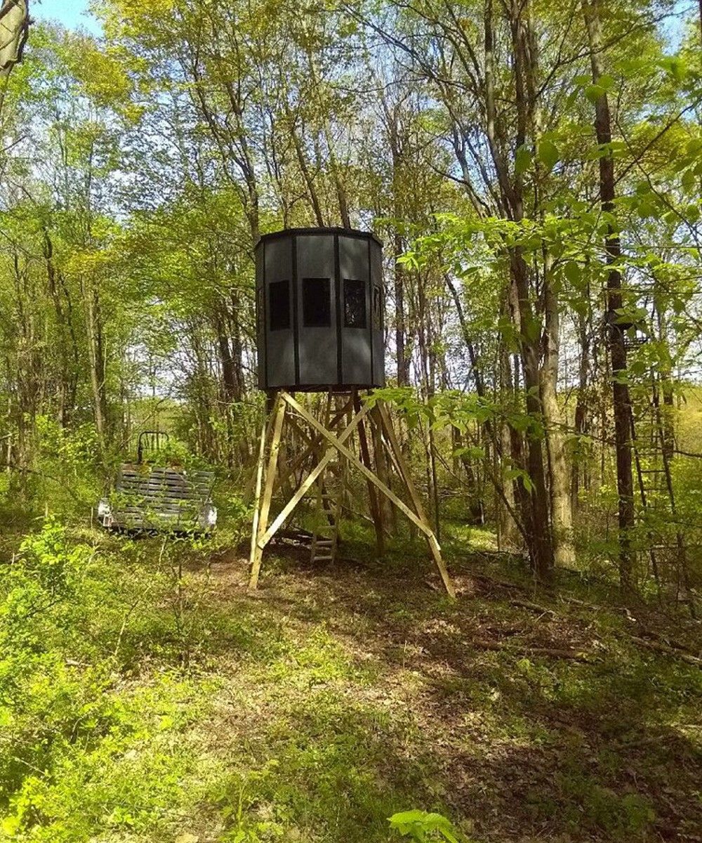 Elevated hunting blind in a forest setting; wooden support structure. Gray structure, green foliage, blue sky.