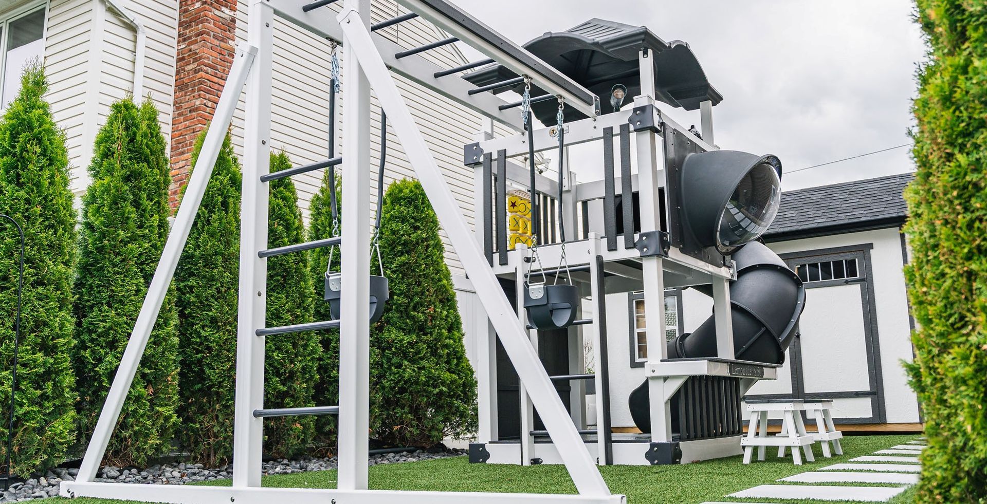 White wooden playset with a slide, monkey bars, and climbing ladder in a yard with green turf and bushes.