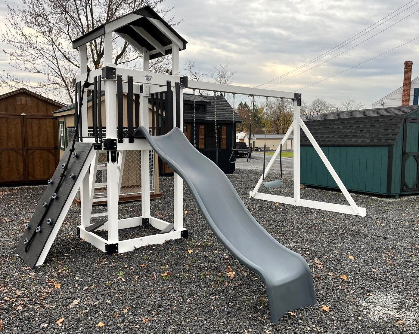 Playground set with slide, climbing wall, swing, and tower in a gravel yard.