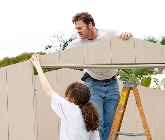 Small shed in a yard, surrounded by a wooden fence, green grass, and plants. Blue sky.