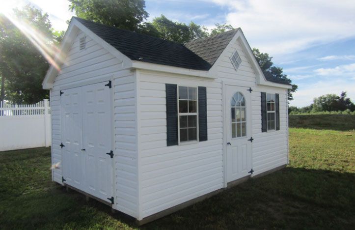 White shed with black shutters and door, in a grassy area, under a blue sky.