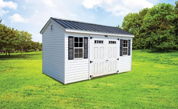White shed with black shutters and roof on a grassy field with trees.