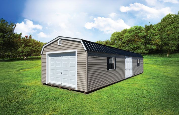 Gray shed with white garage door and black roof on a green lawn with trees and a blue sky.