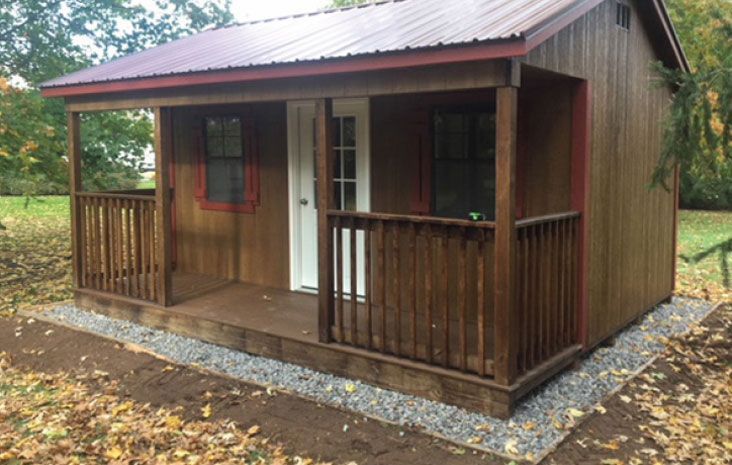 Small brown wooden shed with porch and red trim. Gray gravel borders base, with autumn leaves visible.