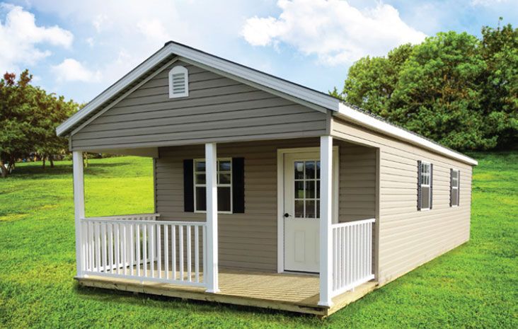 Tan and gray shed with white porch, door, and trim on grassy lawn, blue sky backdrop.