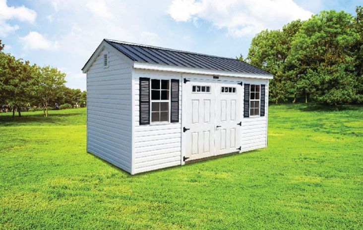 White shed with black roof and shutters in a grassy yard, trees in the background.