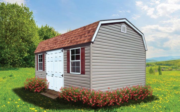 Tan barn-style shed with red roof, white double doors, and pink bushes in a green field.