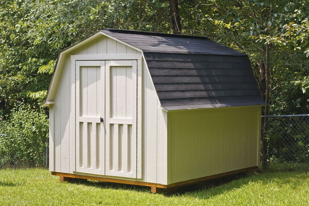 Tan barn-style shed with black roof and double doors in a grassy yard, surrounded by trees.