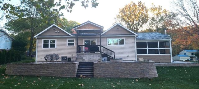 Single-story beige house with front steps, stone retaining wall, and screened porch in a wooded yard.