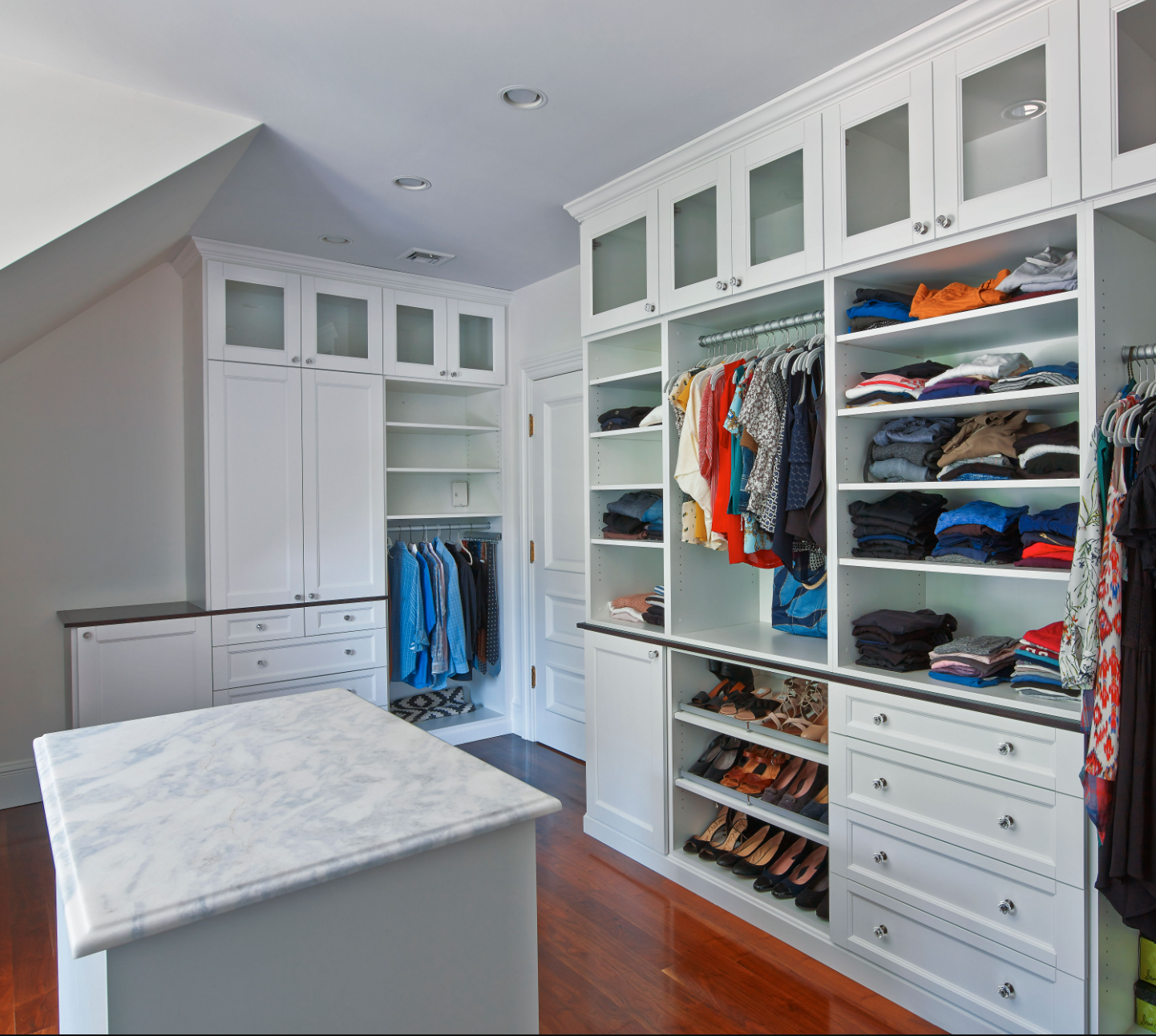 Bright walk-in closet with white built-in shelves, drawers, hanging clothes, and a central island.