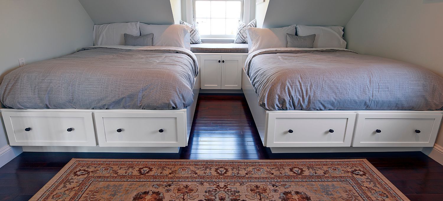 Twin beds with gray bedding in a bright bedroom, with a rug and window between them