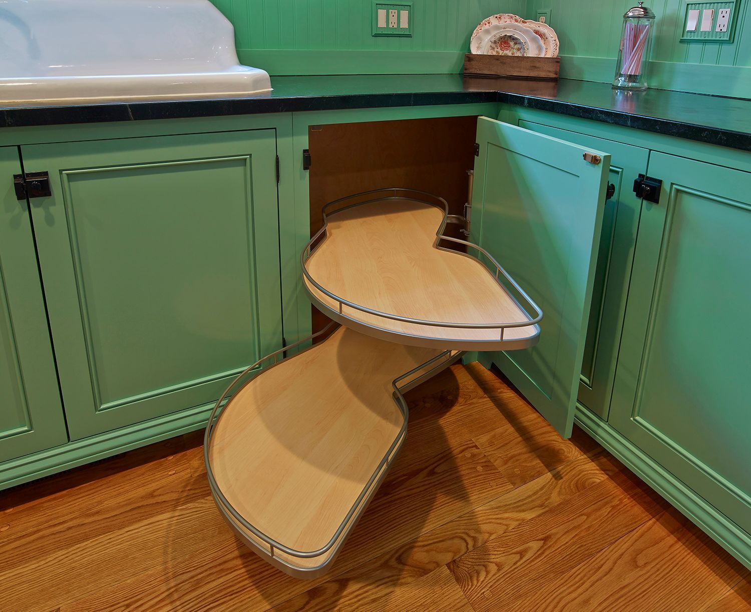 Green kitchen cabinet with pull-out wooden corner shelves open on a wood floor.