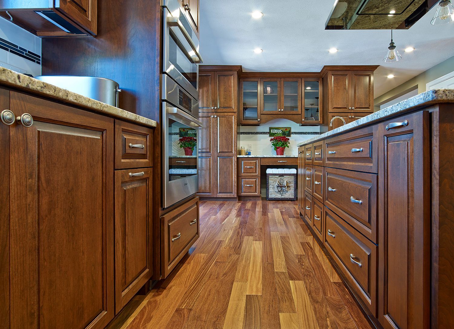 Wooden kitchen interior with cabinets, stainless appliances, and a hardwood floor leading to a distant window.