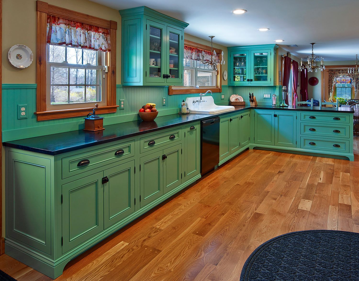 Turquoise kitchen with black countertops, green cabinets, wood floors, and a window over the sink.