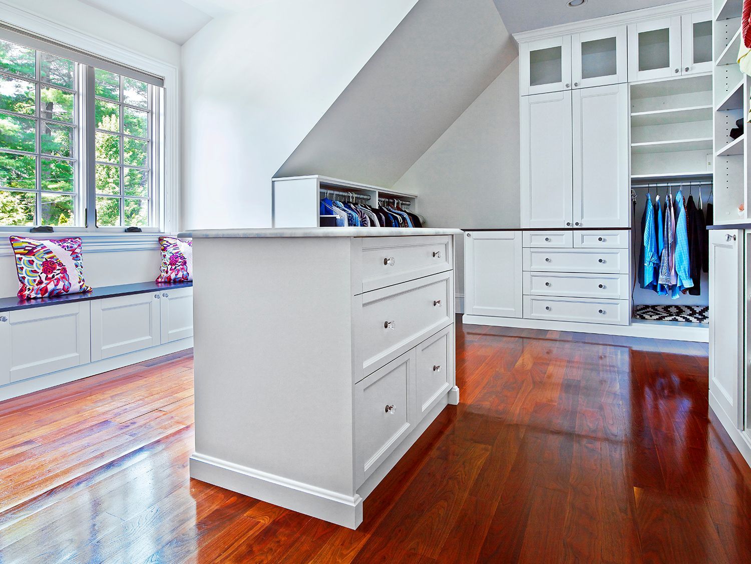 Bright white walk-in closet with drawers, hanging clothes, and hardwood floors beside a window seat.