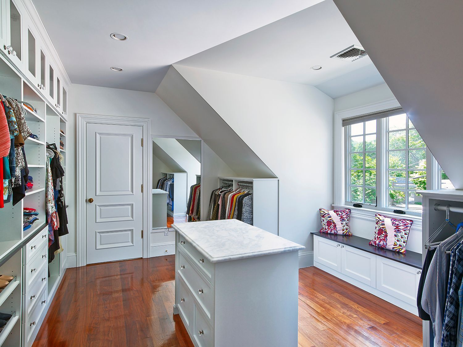 Bright white bedroom with hardwood floors, built-in window seat, and mirrored dresser near slanted ceiling.