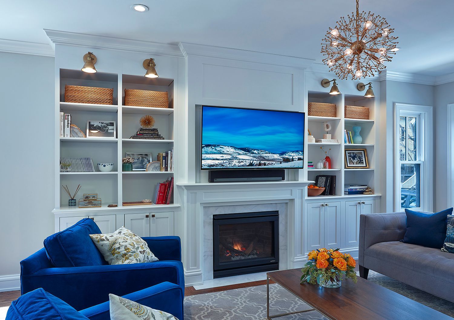 Bright living room with blue chairs, white built-ins, fireplace, and a wall-mounted TV above the mantel
