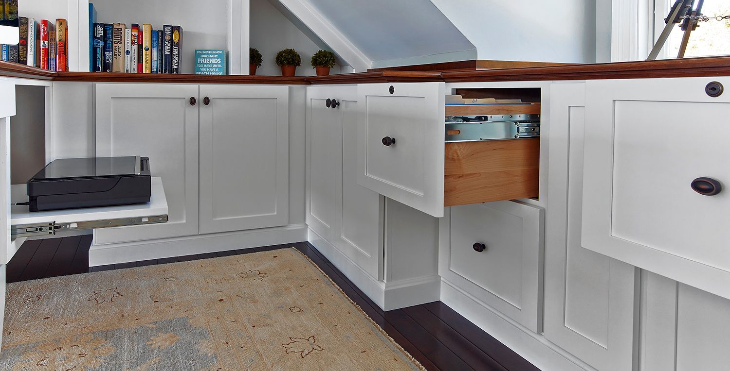 Built-in white cabinets with drawers and shelves along a sloped attic wall, with books and a printer on top.