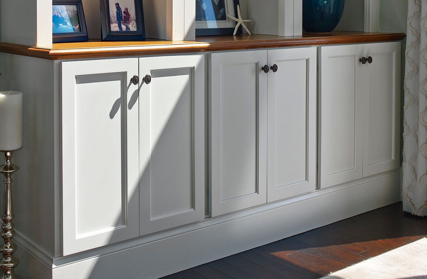 White wooden sideboard with cabinet doors in a sunlit room, topped with framed photos and decor.