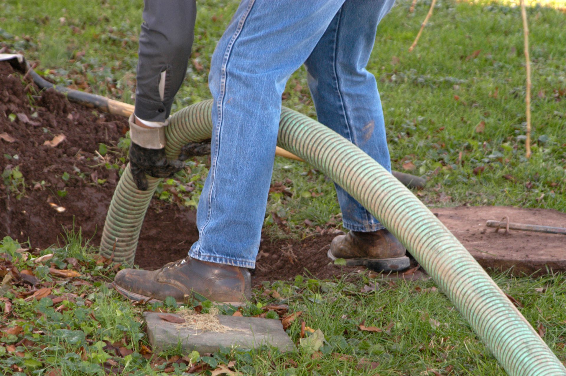 Person in jeans and work boots, holding a large hose near a dirt hole in the grass.