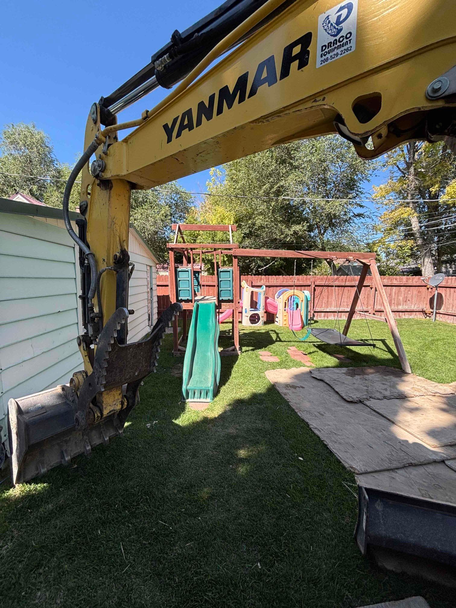 Yellow excavator next to a backyard playground with a slide, swings, and fence.
