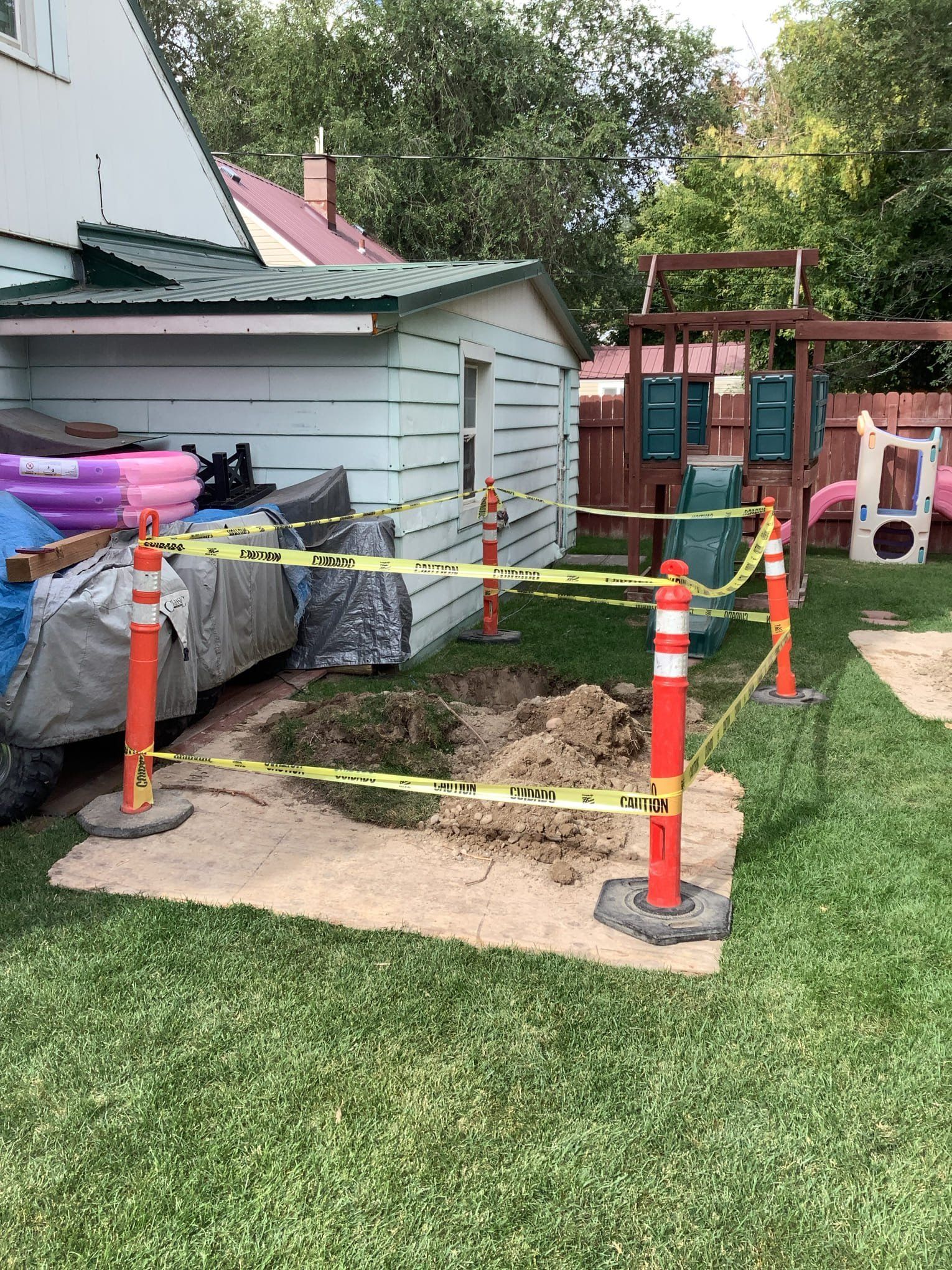 A fenced-off excavated area in a backyard, near a shed and playset.