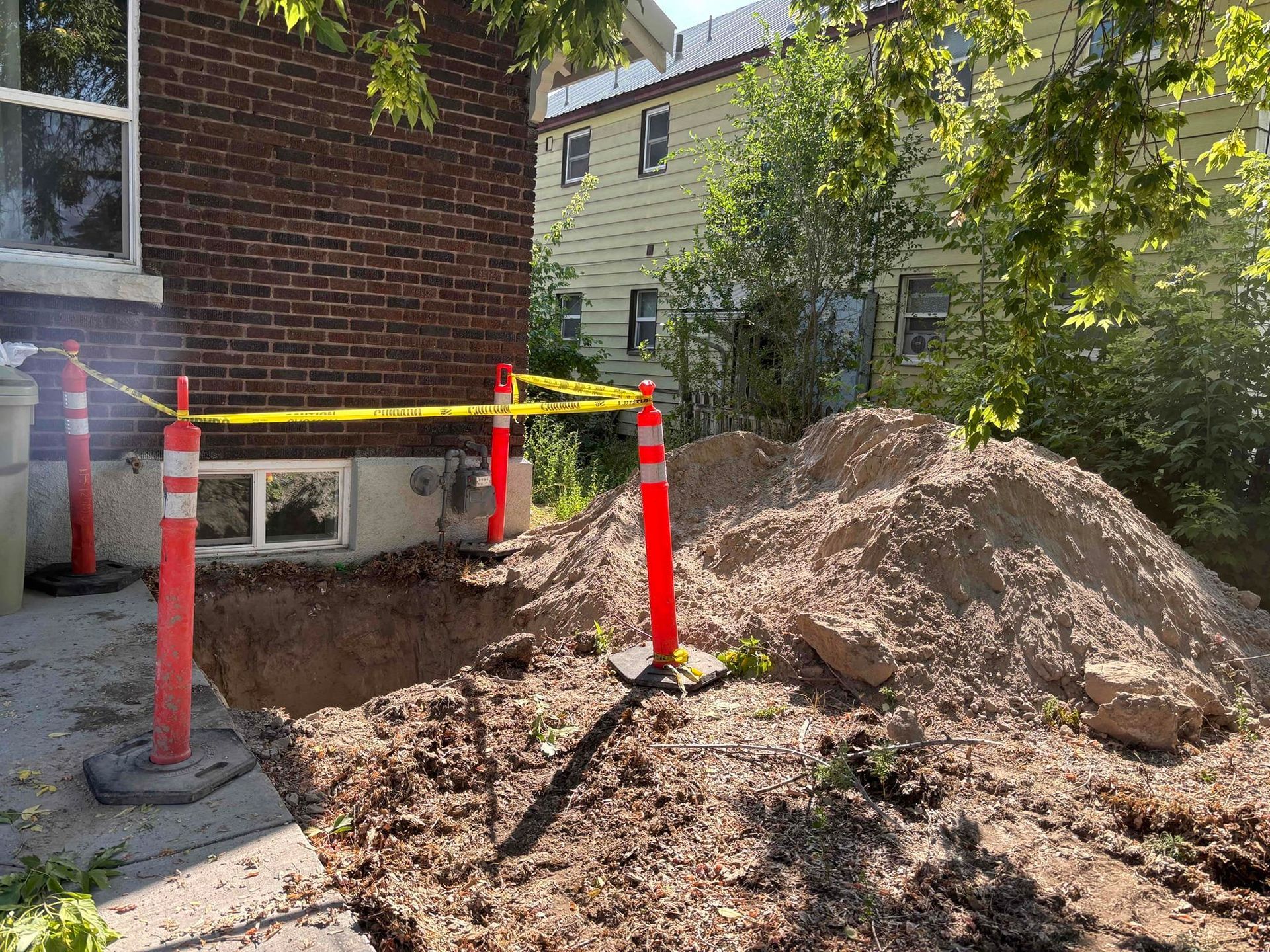 Construction site next to a brick building, with a hole and pile of dirt, orange barriers, and caution tape.