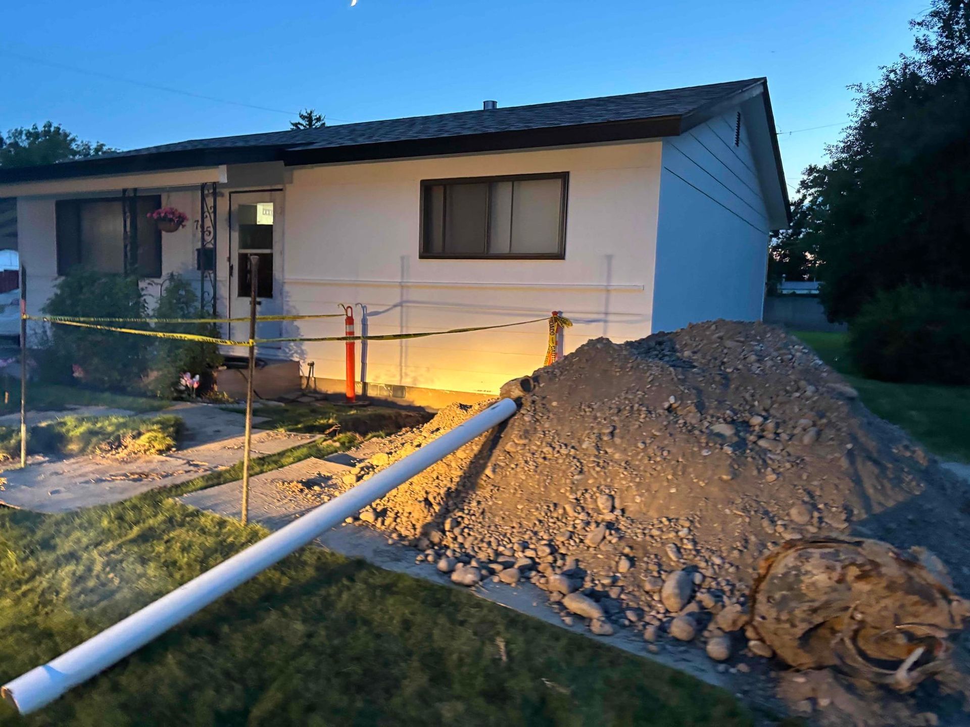 House with construction. Pile of dirt, white pipe, caution tape, and a dim, evening setting.