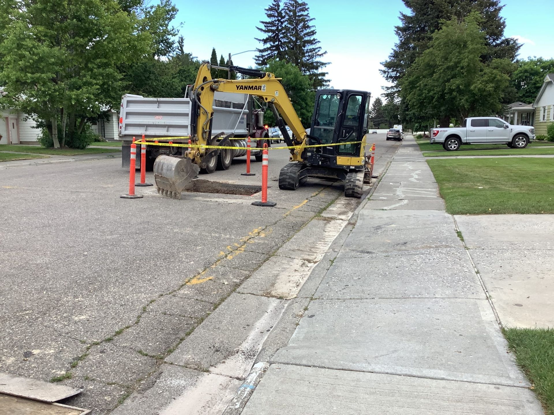 Construction site with an excavator removing concrete from a road next to a sidewalk, with a truck and cones.