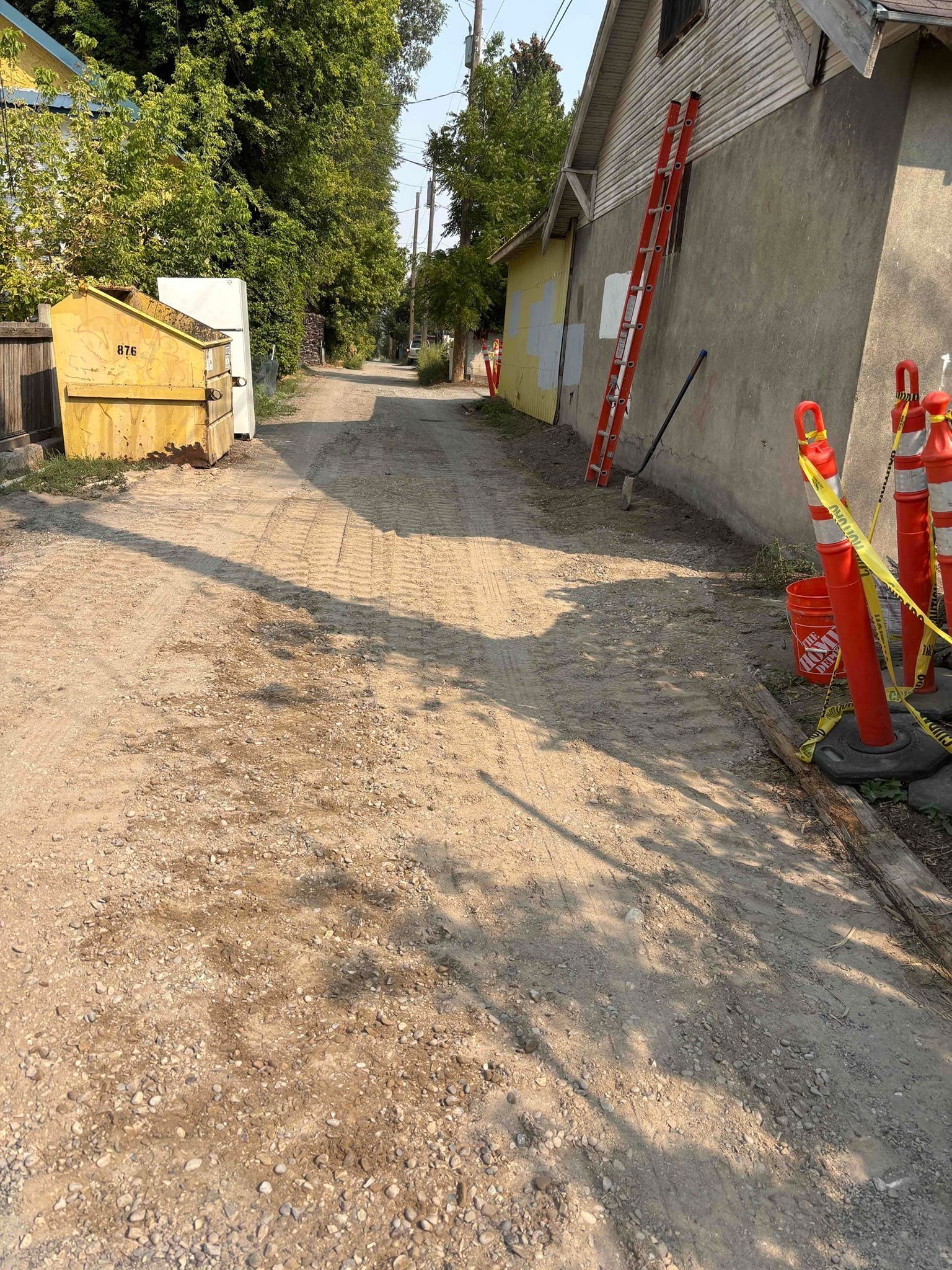 Gravel alleyway with construction cones and a ladder against a building. A dumpster sits at the entrance.