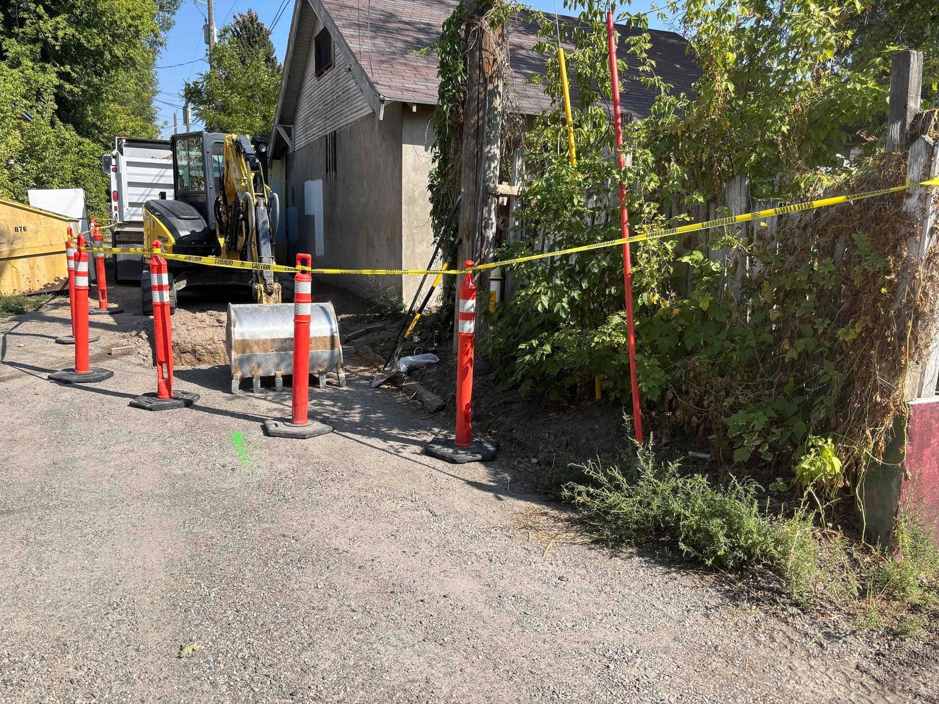 Construction site with mini-excavator, orange cones, caution tape, and a small building.