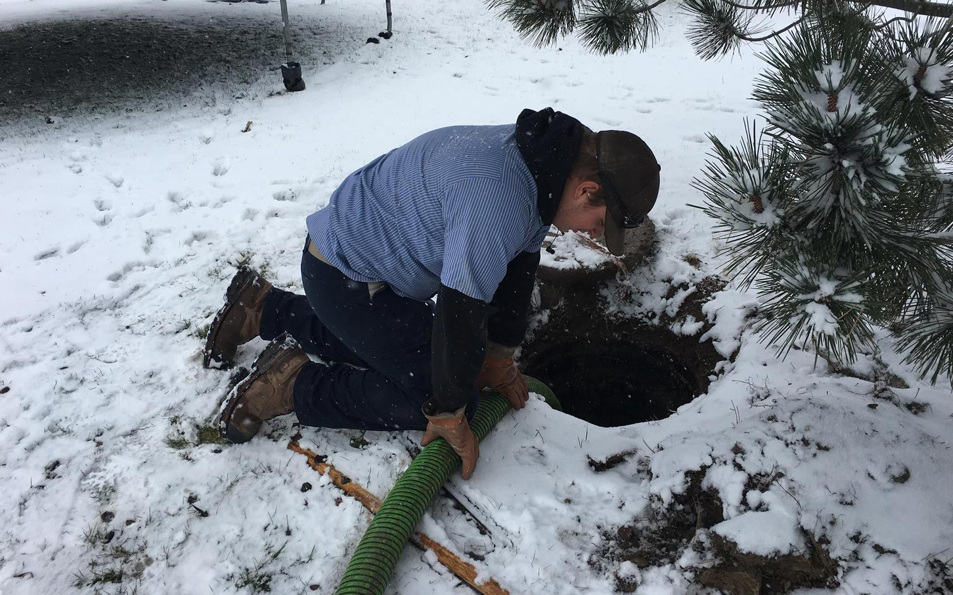 Person kneeling in snow, working on a hole. They're wearing a blue shirt and holding a green hose.