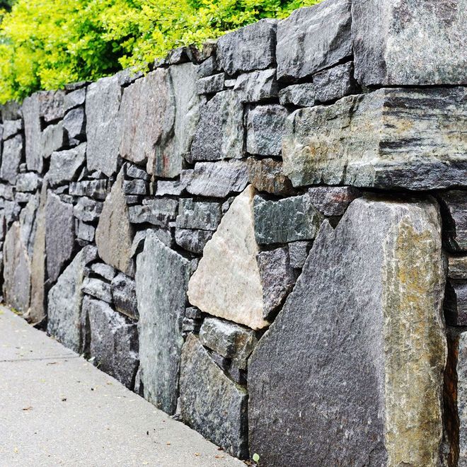Stone wall, gray and tan hues, constructed with various-sized, irregularly-shaped rocks alongside a concrete path.