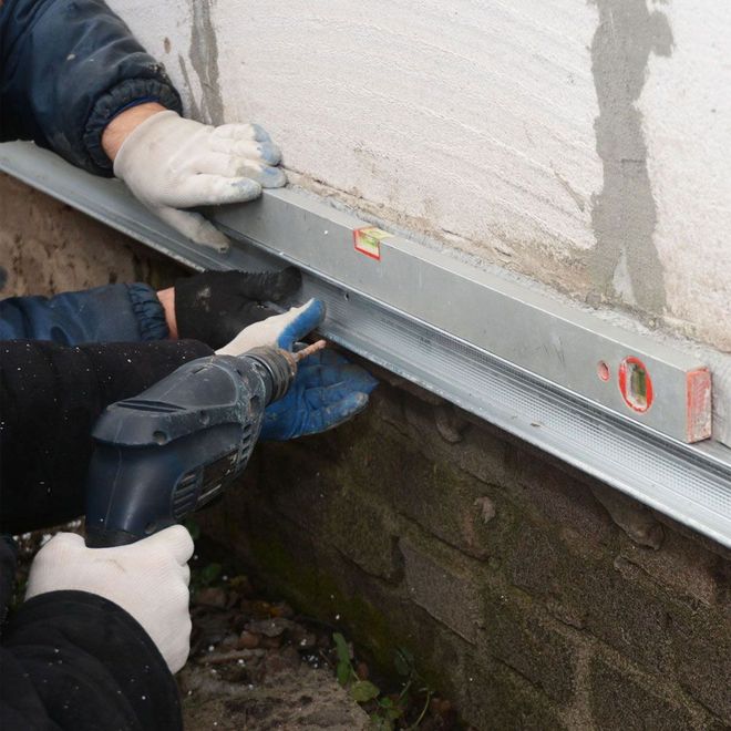 Workers installing metal framing along a concrete wall, using a level and drill.