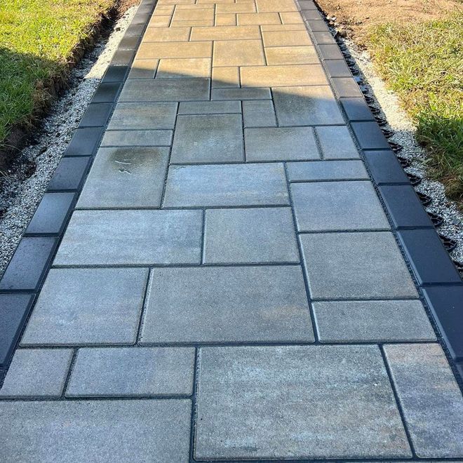 A paved walkway with gray and rectangular stones, bordered by black bricks and gravel, next to green grass.