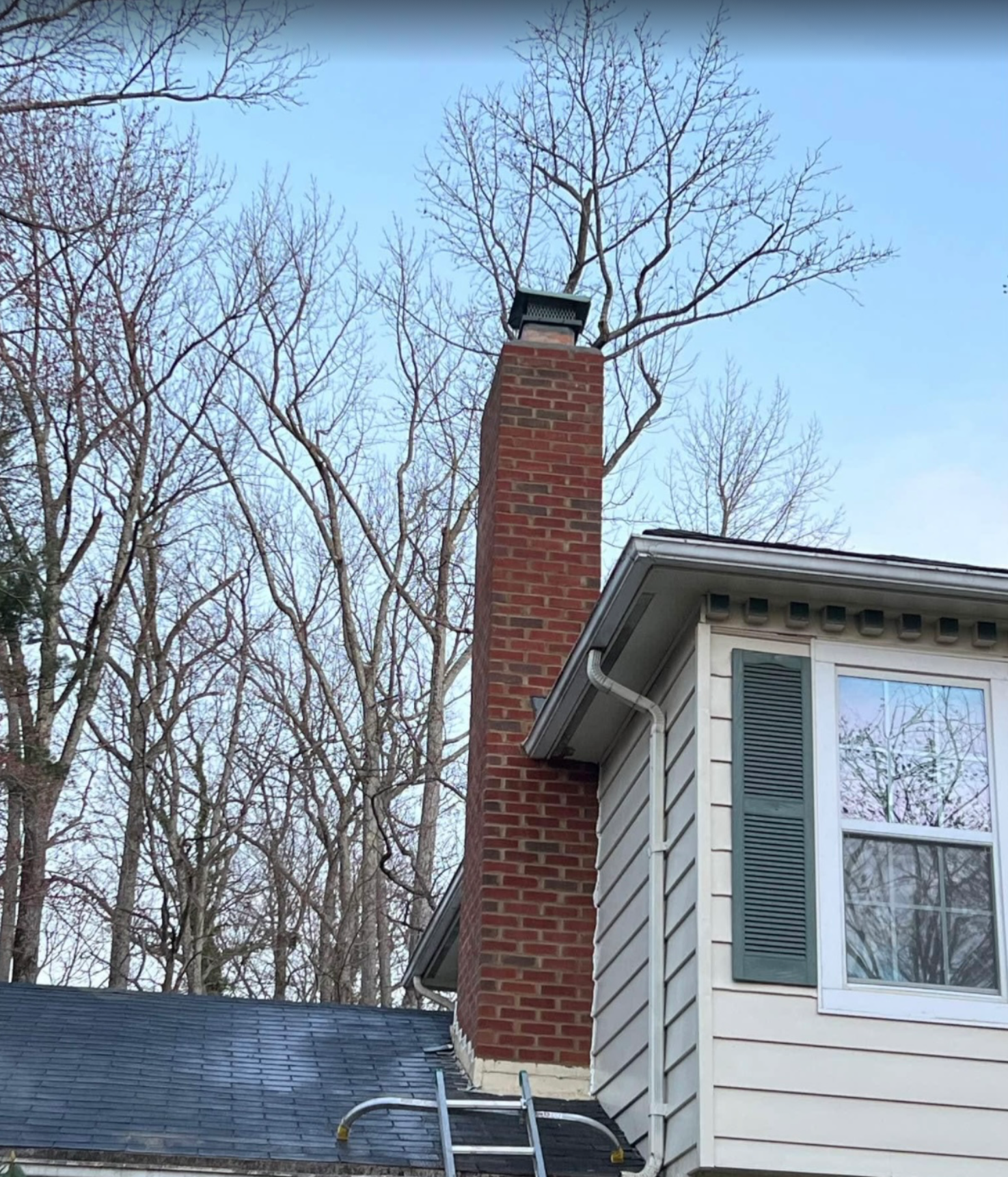 Brick chimney on a house with a ladder and bare trees in the background against a light blue sky.