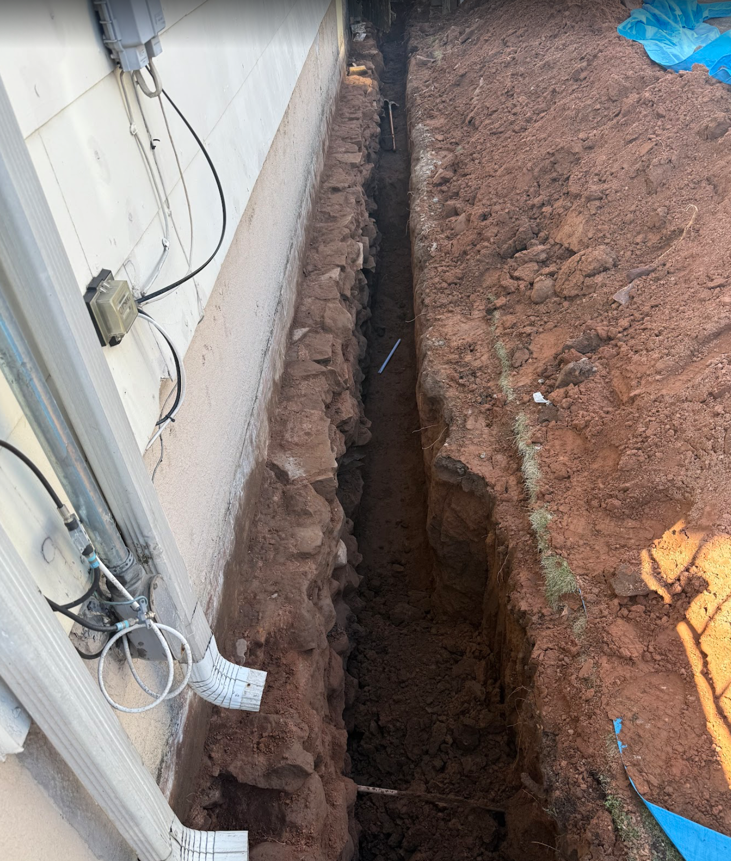 A narrow trench dug next to a building, filled with dirt and debris.