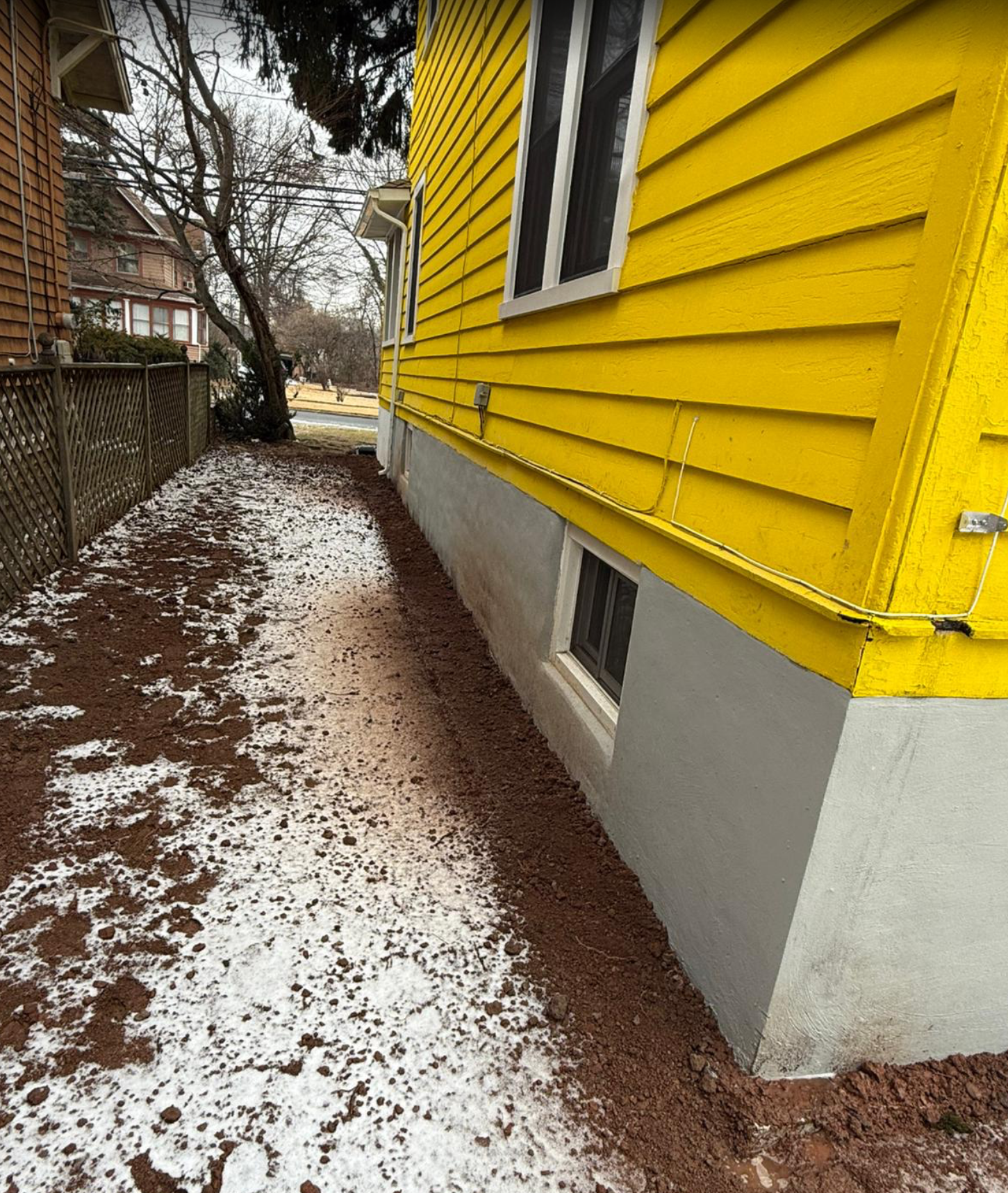 Yellow house with gray foundation, a strip of brown earth and patches of snow along its side.