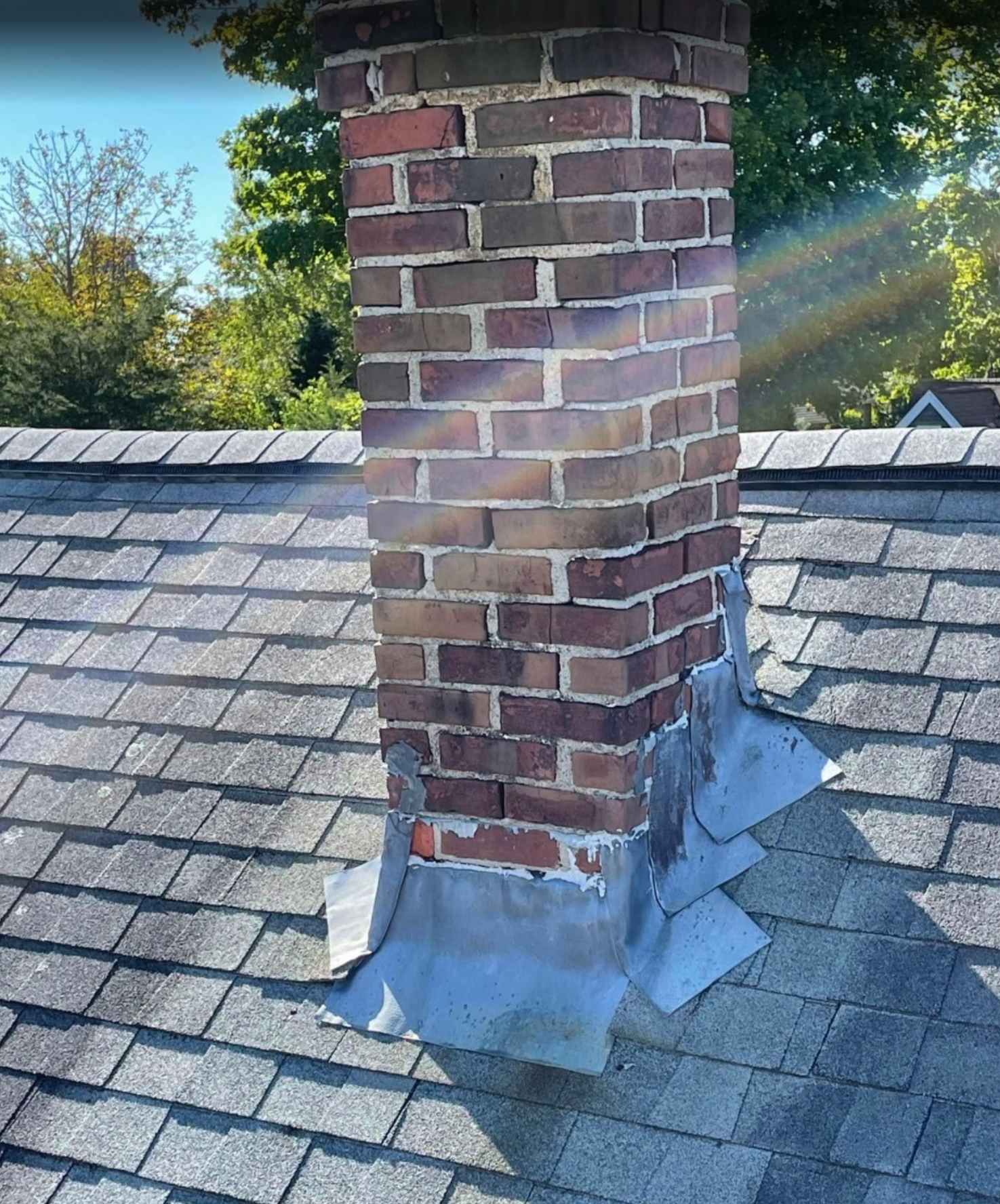 Brick chimney protruding from a shingled roof, with visible flashing and surrounding trees.