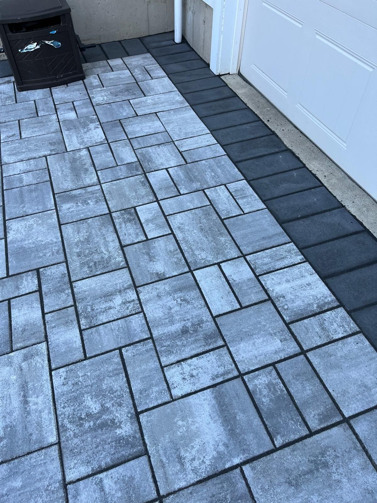 Gray and black paving stones forming a walkway next to a white garage door and a trash bin.