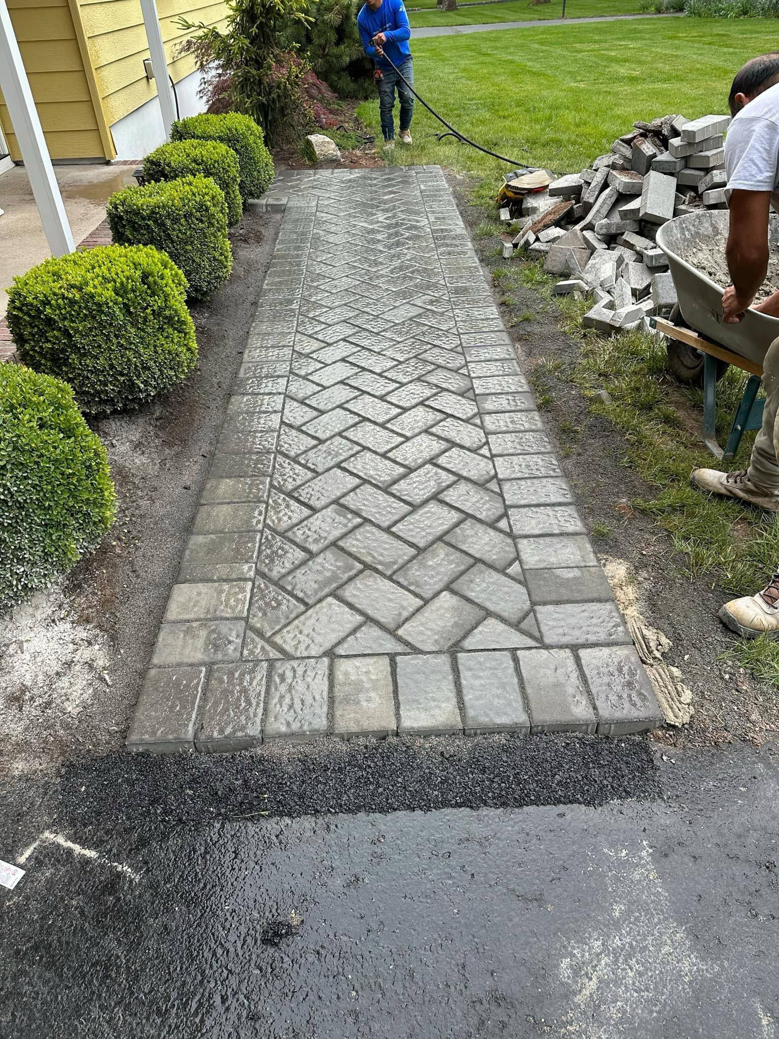 Paver walkway construction: brick pattern, two workers, shrubs, and gravel border in yard.