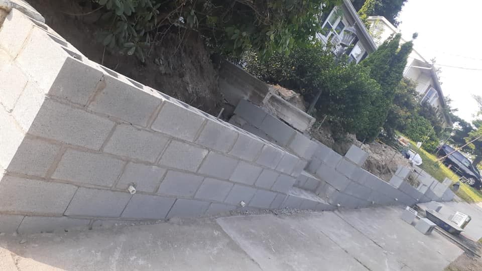 Gray concrete block retaining wall built on a slope, with green vegetation and houses in the background.